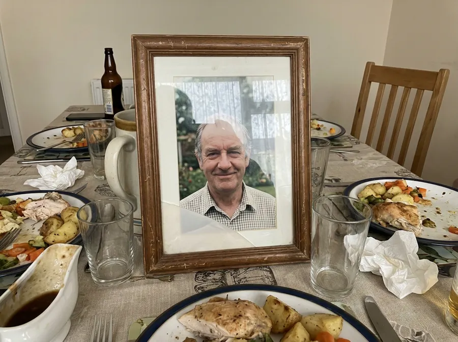 An old framed photograph of a man in his sixties propped on a family dinner table among half-finished roast dinners, with an empty chair pulled out behind it.