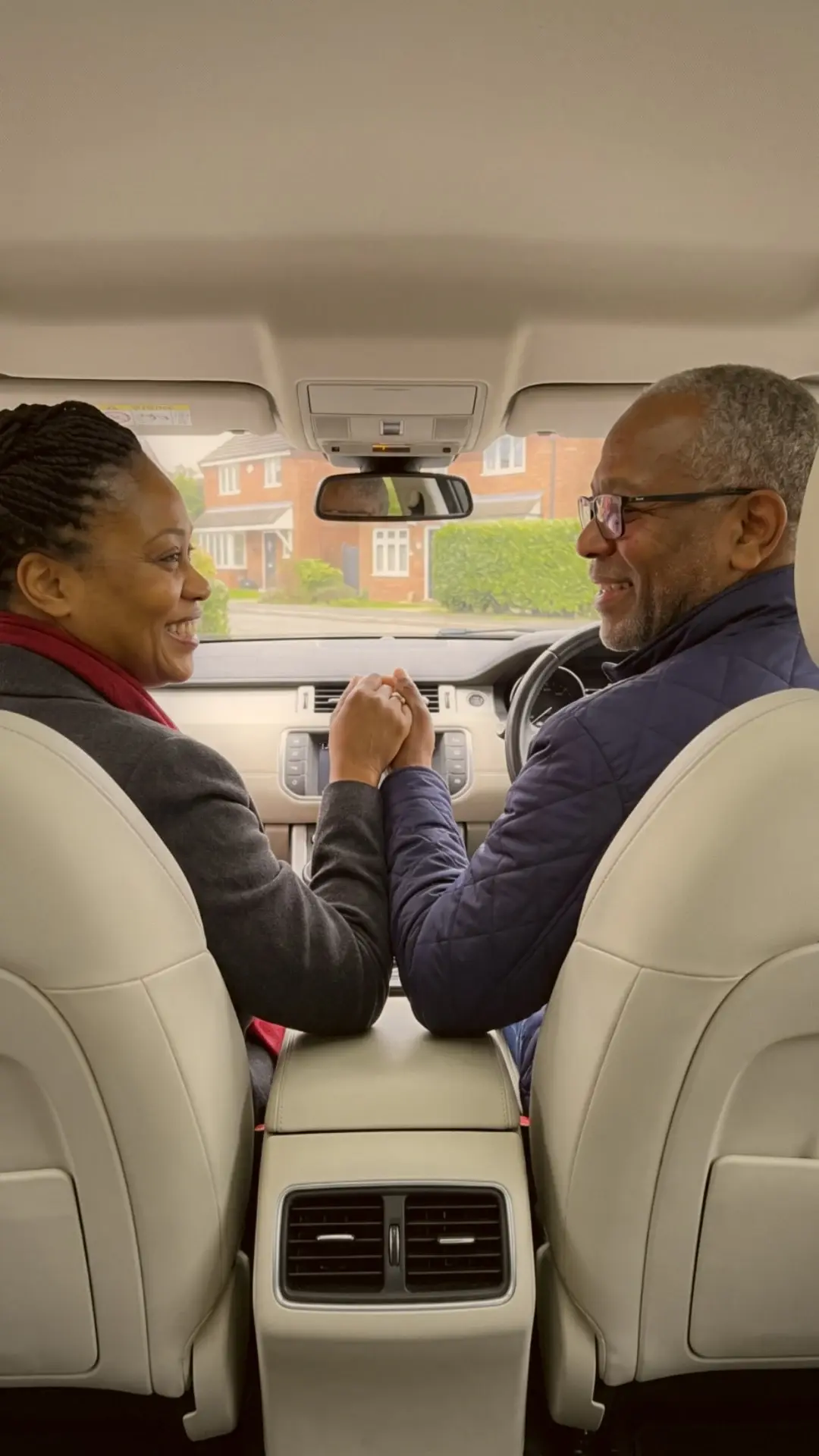 An older couple in the front seats of a car, holding hands across the middle and smiling at each other.