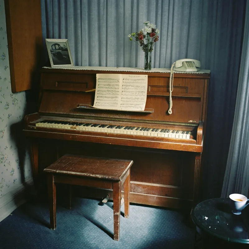 An old upright piano in a quiet front room, sheet music open, a framed photograph and a corded telephone on top, morning light through net curtains.