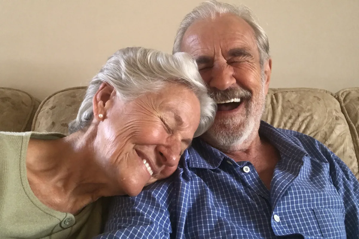 A couple with silver hair laughing together on a sofa at home, her head resting on his shoulder.