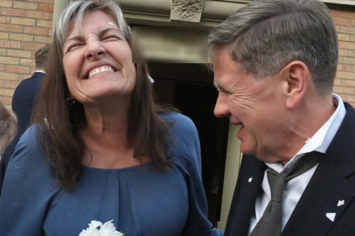 A couple laughing outside a registry office on their wedding day, she in a blue dress holding white flowers.