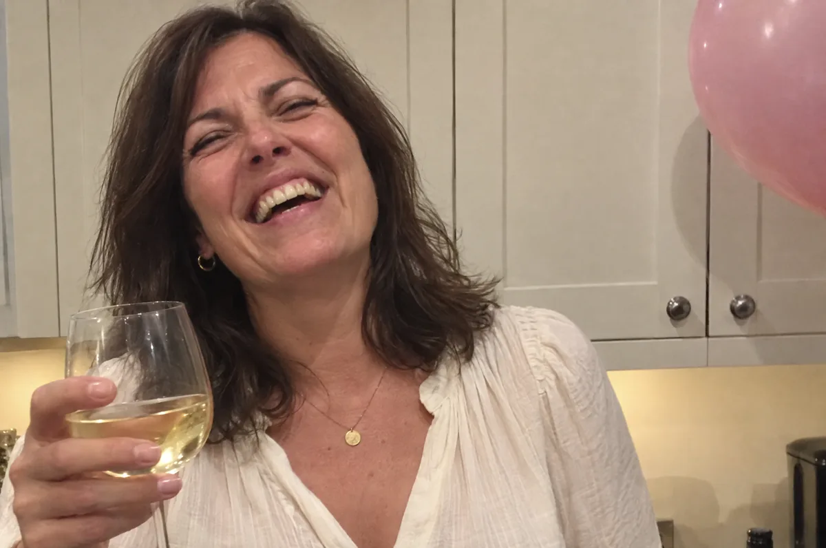 A woman laughing in her kitchen with a pink balloon behind her.